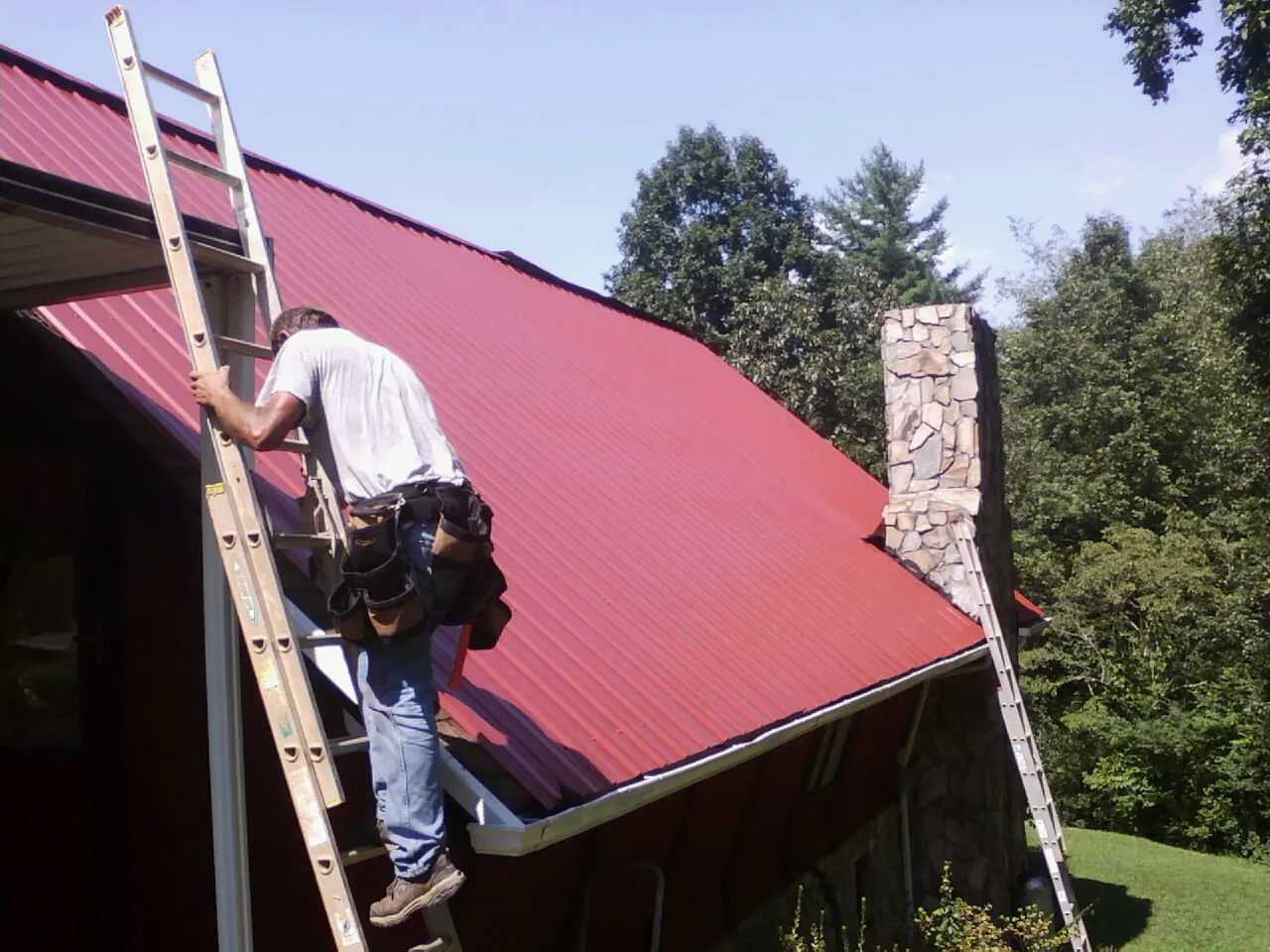 Roofer on a ladder beside a red metal roof and stone chimney — Soffit & Fascia Repair by Roofing Brothers in Grandville